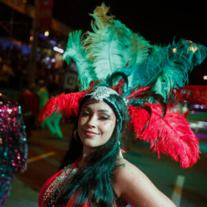 Una mujer con una corona/tocado con grandes plumas verdes y rojas en medio del desfile nocturno