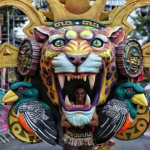 Un artista usando de máscara/botarga la enorme cara de un jaguar ornamentado con una corona estilo música y dos aves trogones, el artista se divisa dentro de la boca rugiente del felino