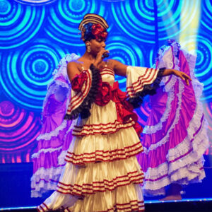 Bailarina cantando en un escenario mientras viste un traje tradicional color crema con detalles rojos, flores y varios volantes. Lleva un tocado en la cabeza y sostiene un micrófono. Al fondo, otra bailarina sostiene una gran falda roja y blanca mientras las luces azules y moradas iluminan el escenario.