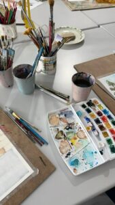 Una mesa llena de implementos para pintar, acualeras, vasos y lienzos.