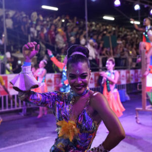 En medio de un desfile, una bailarina saluda a la cámara con una sonrisa.